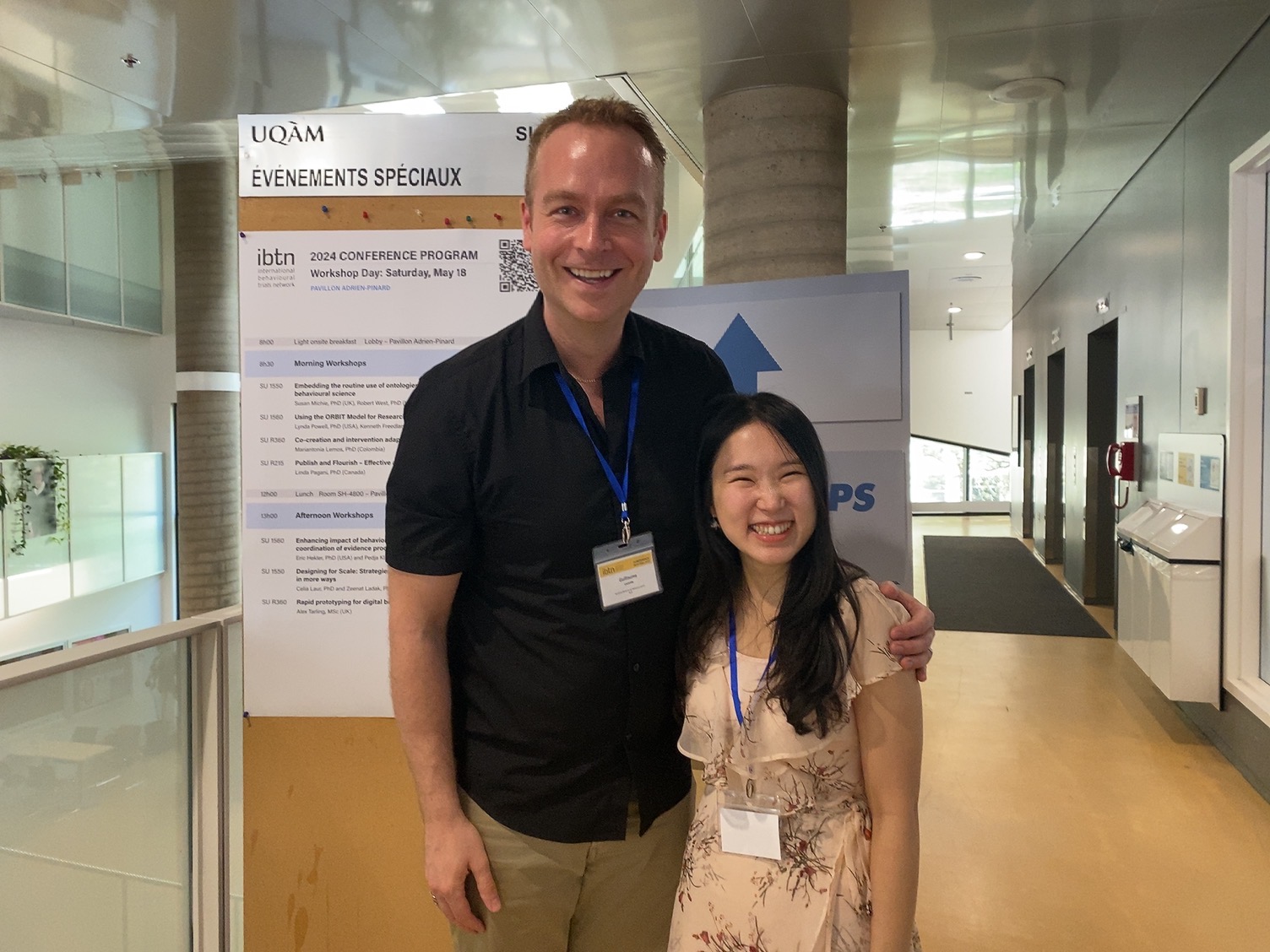 Two people smiling together at a conference in front of a workshop program board