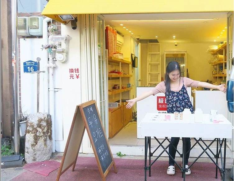 A person standing with arms open behind a small table in front of a shop entrance with a yellow awning