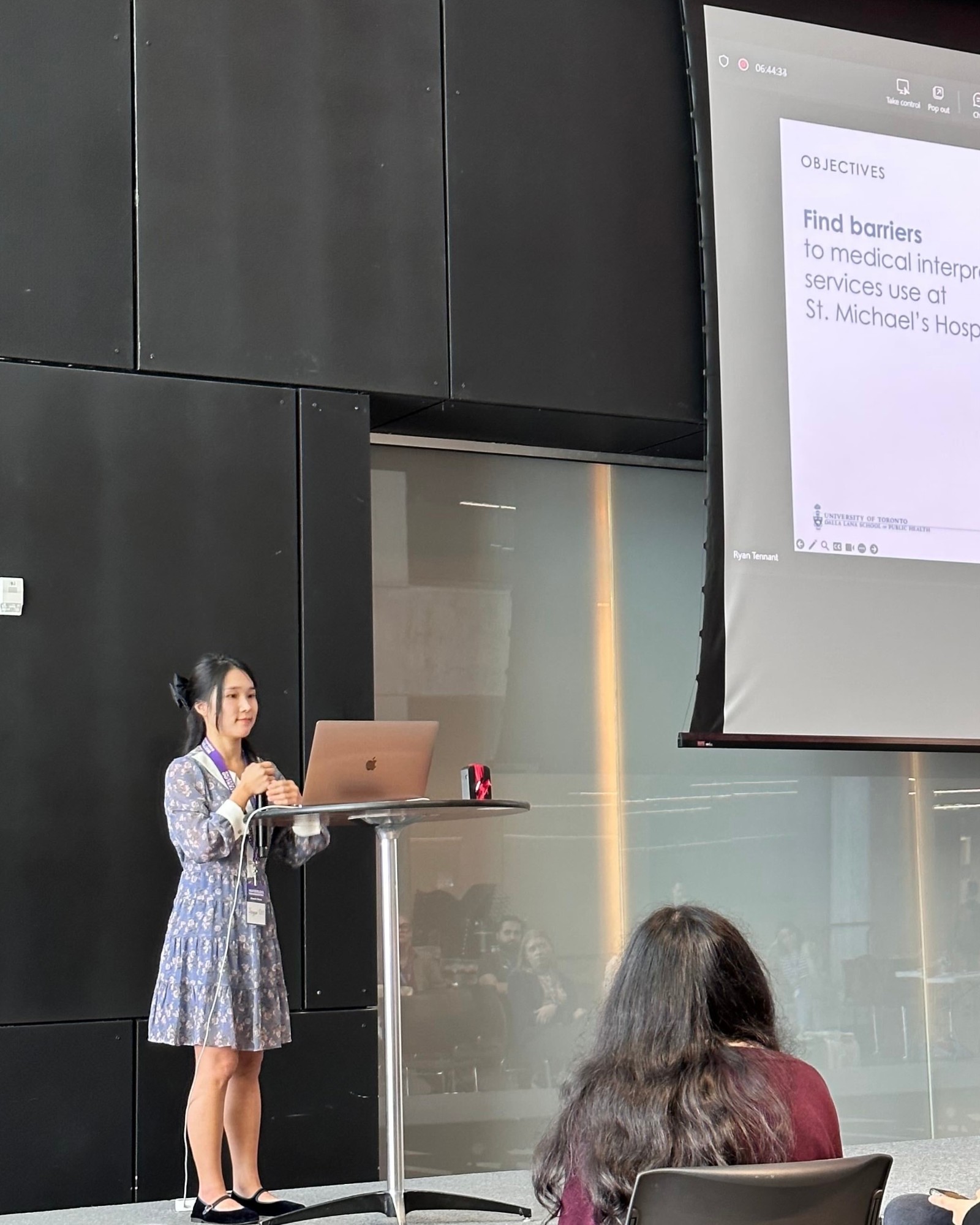 A person giving a presentation at a standing table with a laptop, next to a projected slide