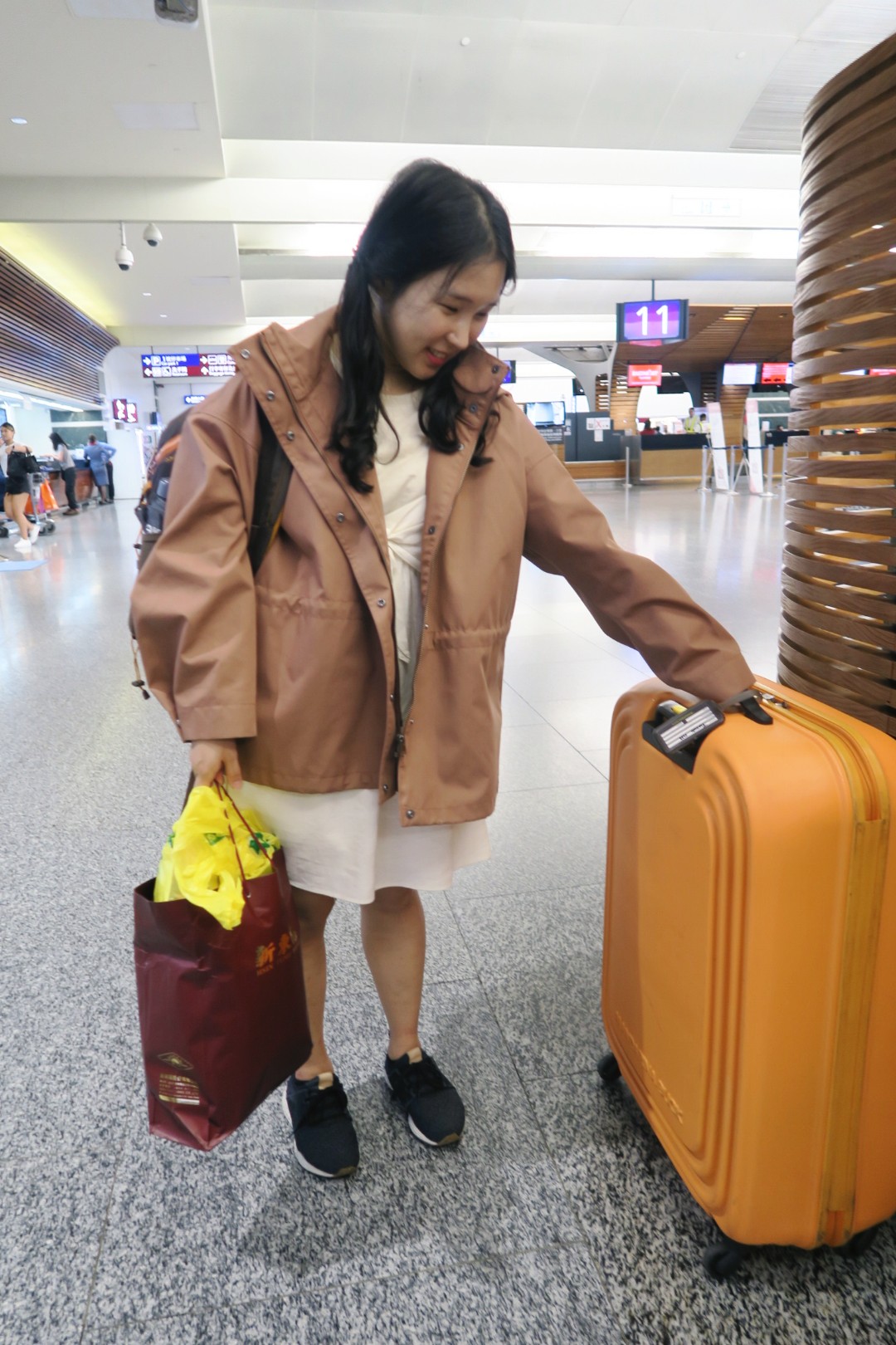 A person in a tan jacket holding an orange suitcase and a shopping bag in an airport