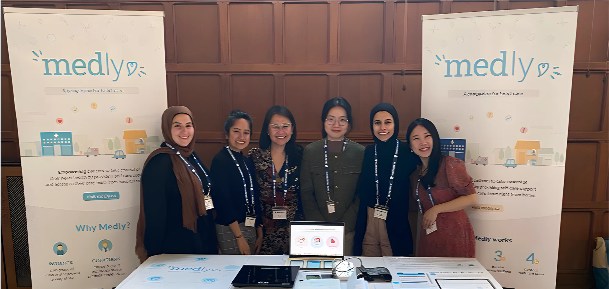 Six people smiling behind a Medly booth table, between two Medly banners at a conference