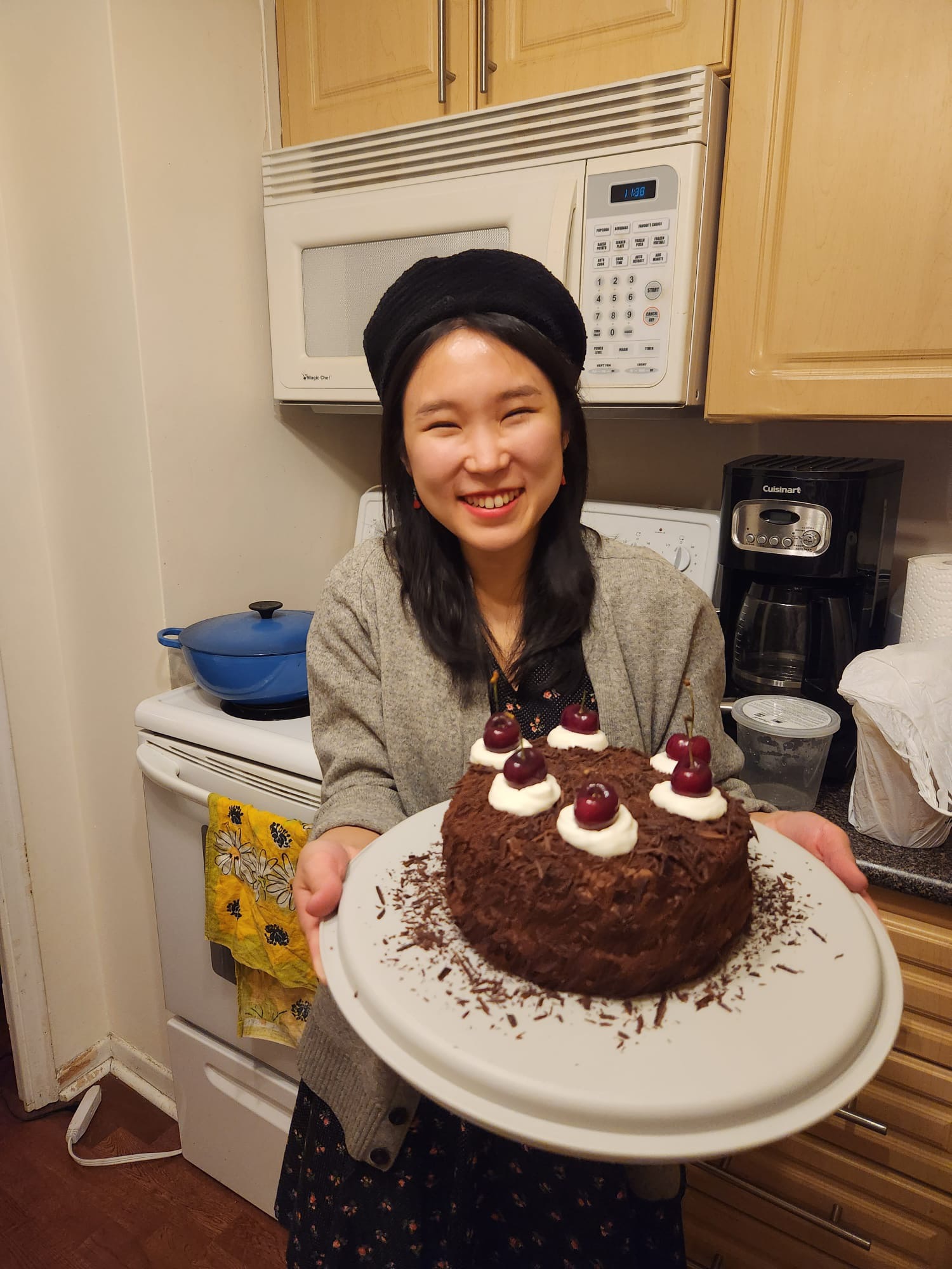 A person in a black beret smiling and holding a chocolate cake with cherries on a plate in a kitchen