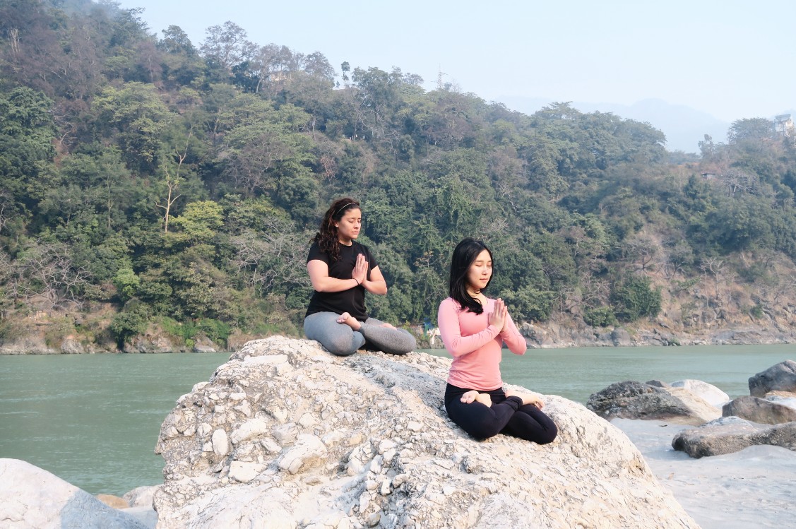 Two people meditating with hands in prayer position on a large rock beside a river