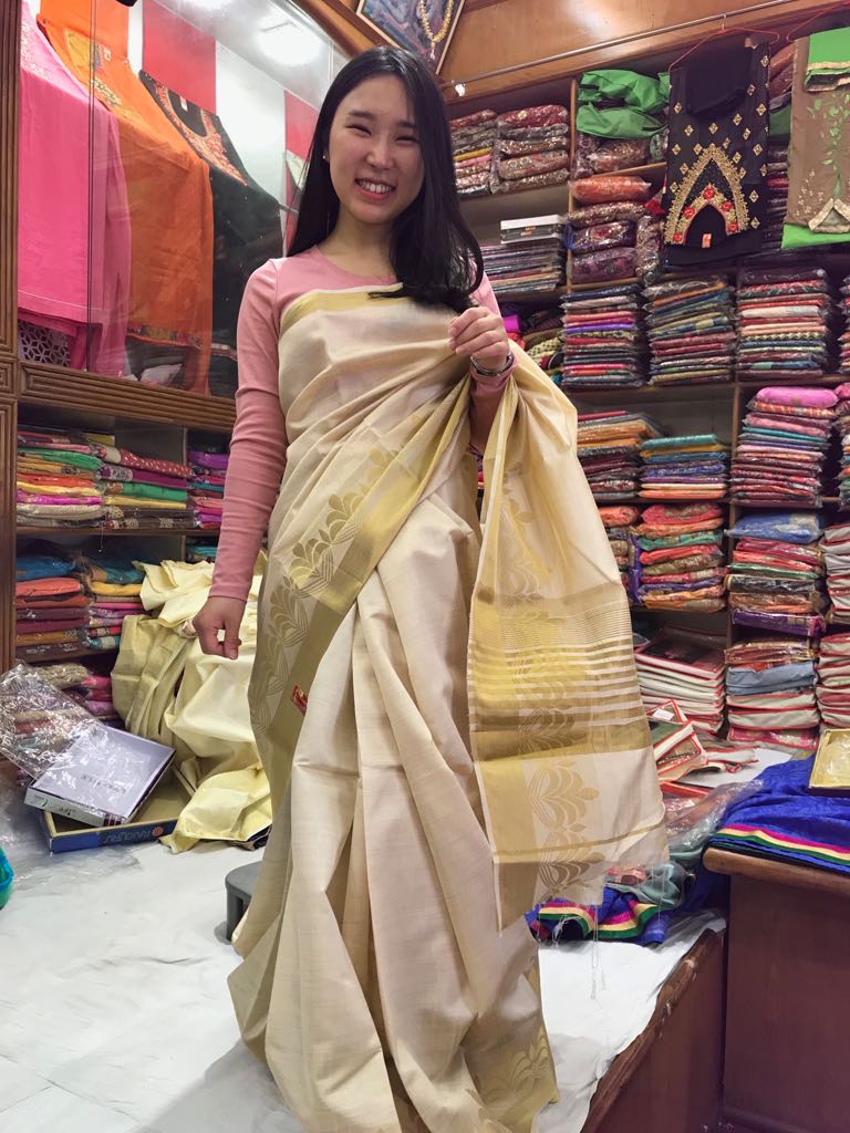 A person smiling in a cream and gold sari inside a fabric shop